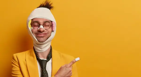 Uomo con benda in testa e cerotto sul dito indica come viene pagato l'infortunio, con un sorriso ironico.