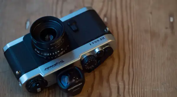 A silver and black Voiglander Bessa L camera with a lens attached and a separate 28mm lens nearby, resting on a wooden surface.