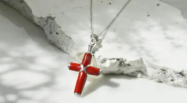 Collar con colgante de cruz de coral rojo y plata, sobre una superficie texturizada de piedra blanca.