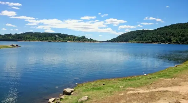 Un día soleado en la ruta del agua La Jarosa, con un embalse azul rodeado de colinas verdes y un cielo con nubes blancas.