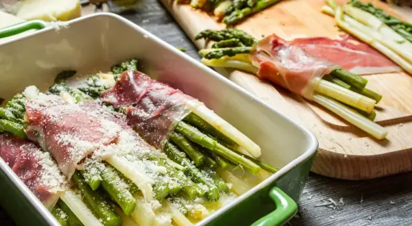 Zielone i białe szparagi w szynce na zimno, posypane parmezanem, gotowe do pieczenia.