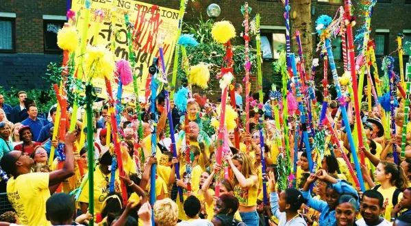 Gente celebra en un desfile, levantando coloridos bastones adornados con pompones y cintas. Un cartel dice "POLE-O-NG STATION". Parece una fiesta, quizás un bank holiday.