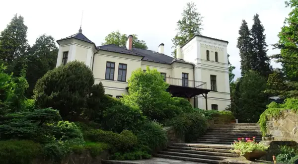 Ein hellgelbes Herrenhaus mit Turm und Balkon, umgeben von üppigem Grün und einer Steintreppe, in Opava, Tschechien.
