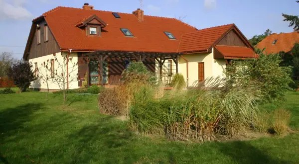 Dom z czerwonym dachem i naczółkami, otoczony zielonym trawnikiem i ozdobnymi trawami.