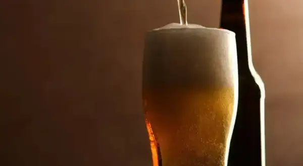 Birra pils che si versa in un bicchiere, con una bottiglia sullo sfondo. Schiuma abbondante e colore dorato invitante.