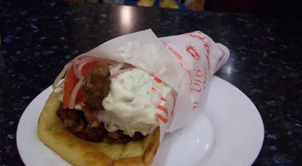 Pyszna pita grecka z mięsem, warzywami i sosami, zawinięta w papier z greckimi napisami.