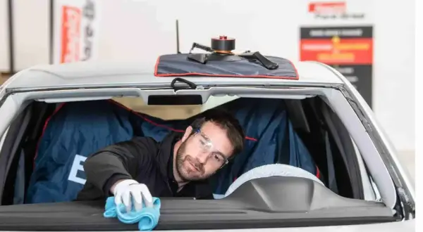 Un technicien porte des lunettes de protection et utilise un chiffon bleu pour nettoyer l'intérieur du pare-brise d'une voiture.