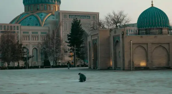 Samotny podróżnik klęczy na pustym placu przed meczetem z turkusową kopułą.