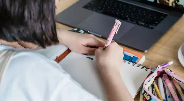 Dziewczynka pisze w zeszycie, obok leży laptop i piórnik z kredkami.