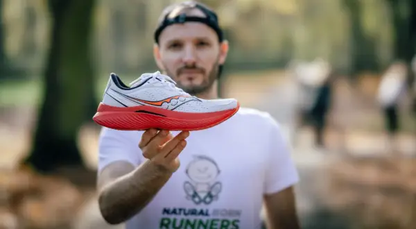 Mężczyzna w koszulce "Natural Born Runners" prezentuje biało-czerwony but biegowy Saucony. Saucony to znana firma z tradycjami.