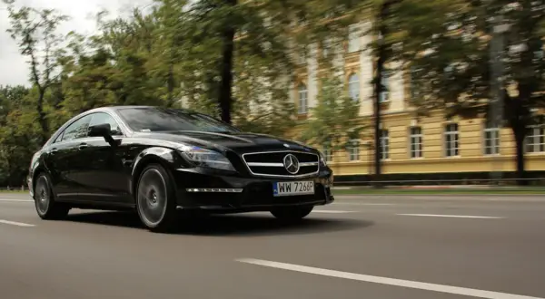 Czarny Mercedes CLS 63 AMG Coupe pędzi ulicą, mijając zabytkową kamienicę i drzewa.