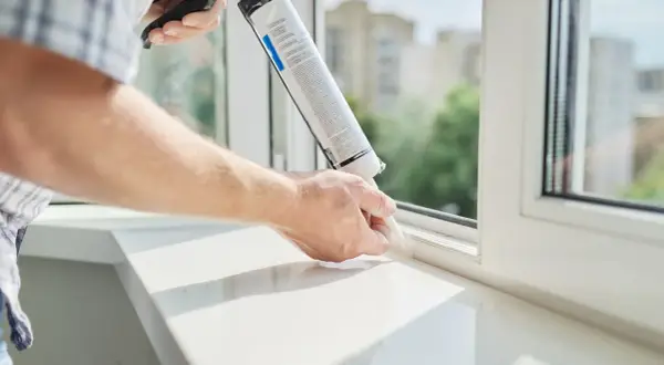 Mężczyzna aplikuje masę uszczelniającą, pokazując jak zamontować parapet wewnętrzny. Biały parapet i okno.