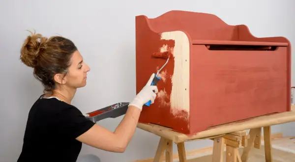 Mujer aplica pintura para aglomerado a un baúl de madera con un rodillo.