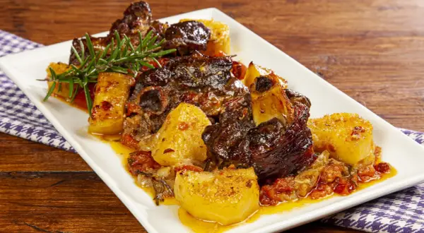 Un saporito agnello con patate, cotto lentamente con pomodorini e rosmarino, servito su un piatto bianco.