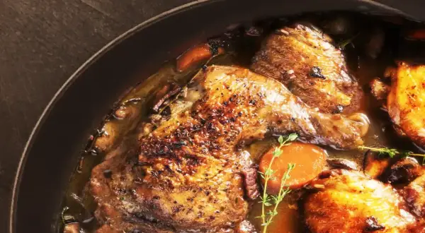 Un plat de coq au vin mijoté, avec des morceaux de poulet dorés, des carottes, des champignons et des herbes dans une sauce riche.