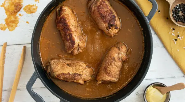 Herzhafte schweinerouladen nach omas rezept in dunkler Soße, bereit zum Servieren. Daneben Senf, Salz und Pfeffer.