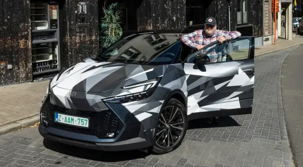 Man lutar sig mot en Toyota C-HR Hybrid i kamouflagemönster. Bilen är parkerad på en kullerstensgata framför en byggnad med en automat.