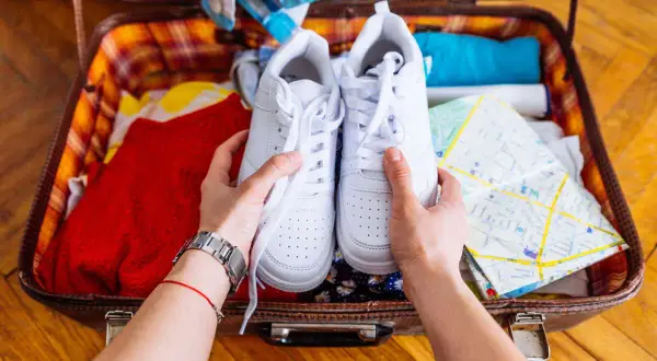 Comment ranger des chaussures dans une valise : une paire de baskets blanches est tenue au-dessus d'une valise ouverte remplie de vêtements et d'une carte.
