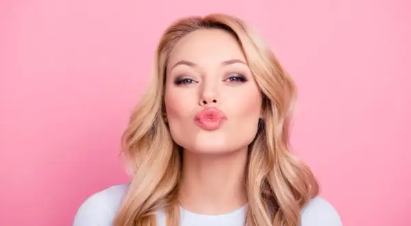 Donna con capelli biondi e trucco labbra naturale, che manda un bacio su sfondo rosa.
