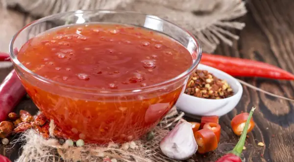 En skål med söt chili sås omgiven av färska och torkade chilifrukter, vitlök och pepparkorn.