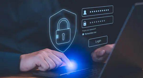 Dłoń dotyka ekranu laptopa z interfejsem logowania. Ikona tarczy z kłódką symbolizuje bezpieczeństwo, a obok pola na login i hasło znajduje się przycisk "Login". To nowoczesny menedżer haseł.