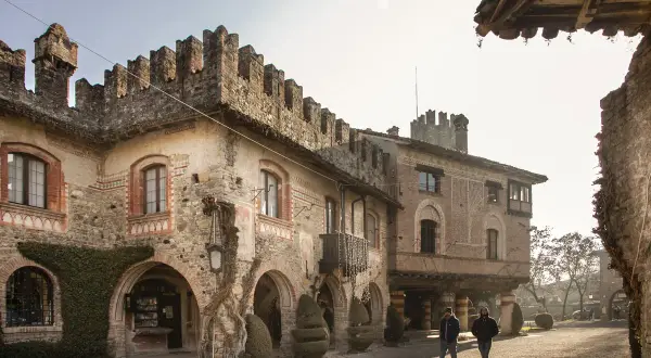 Antico borgo Visconti a Piacenza, con torri merlate e archi in pietra. Due uomini passeggiano nel cortile illuminato dal sole.