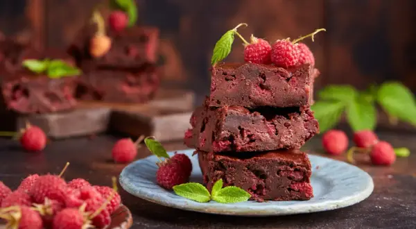 Czekoladowe brownie z malinami, ułożone na talerzyku, ozdobione świeżymi malinami i listkami mięty.