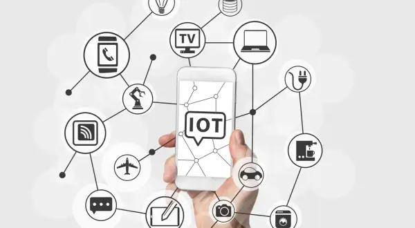 Dłoń trzyma smartfon z napisem "IOT co to". Wokół widnieją ikony urządzeń połączonych siecią: żarówka, TV, laptop, ekspres do kawy, pralka, samochód, aparat, samolot, robot, smartfon, router i chmura danych.