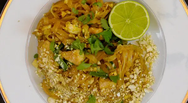 Talerz z Pad Thai, posypany orzeszkami ziemnymi, z połówką limonki i kolendrą.