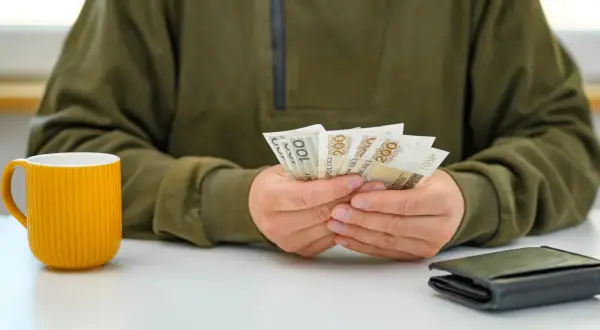 Osoba liczy banknoty 200 zł, symbolizujące podwyżki dla pracowników samorządowych. Obok stoi żółty kubek i portfel.