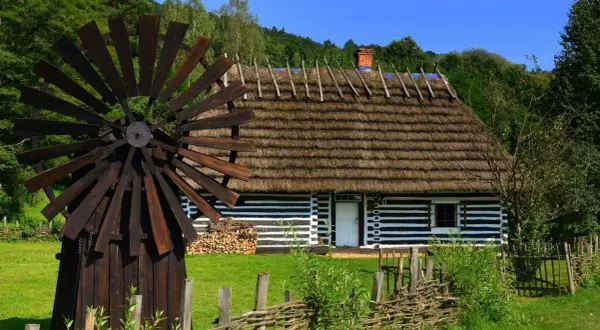 Drewniany wiatrak i tradycyjny dom z krytym strzechą dachem na tle zieleni.