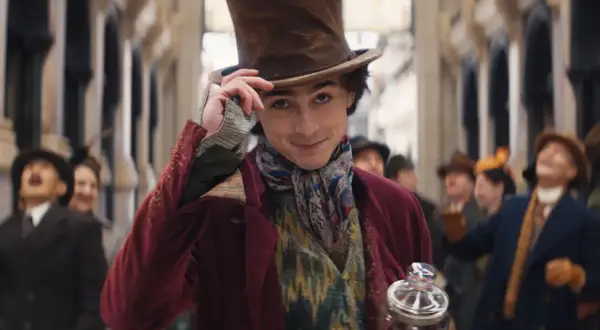 Timothée Chalamet w filmach, jako młody Willy Wonka, w cylindrze i barwnym stroju, z uśmiechem pozdrawia tłum.