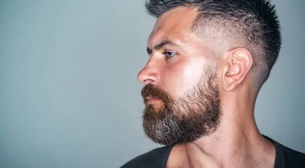 Mann mit Bart und tiefer fasson haarschnitt, im Profil auf grauem Hintergrund.