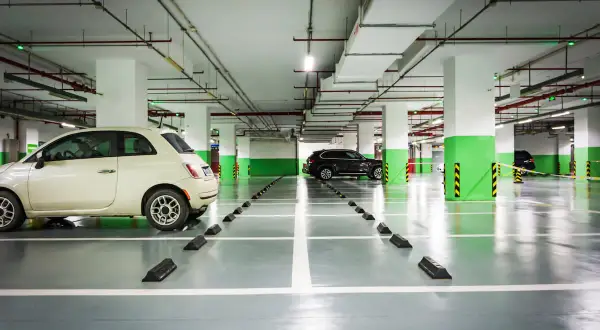 Jasny samochód i ciemny SUV stoją na podziemnym parkingu. Widać wyraźnie wyznaczoną szerokość miejsca parkingowego, z odbojnikami na każdym stanowisku.