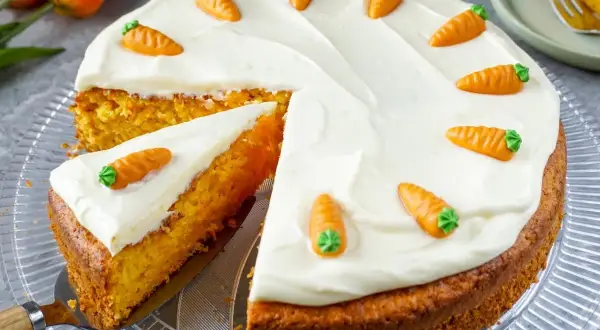 Saftiger Karottenkuchen mit cremiger Glasur und kleinen Marzipan-Karotten. Ein Stück ist bereits angeschnitten.