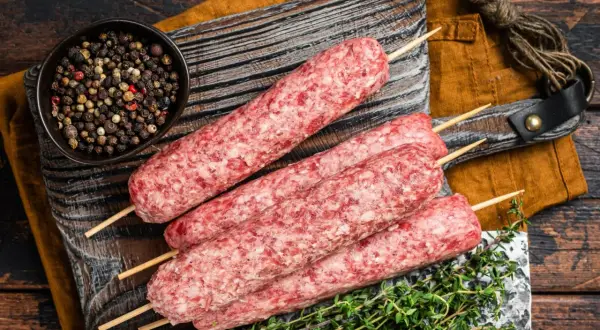 Surowe kofty wołowe na patyczkach, gotowe do grillowania, obok miseczki z pieprzem i gałązki tymianku.