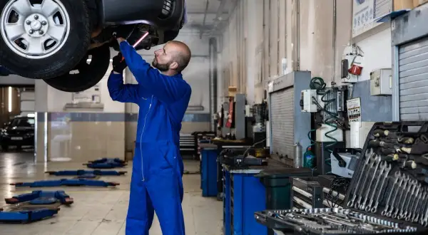 Mécanicien en bleu travaillant sous une voiture soulevée, outil à la main. Il pourrait s'agir d'une étape pour comment nettoyer un fap démonté.