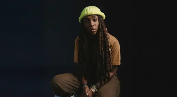 Jackie Hill Perry, mit Dreadlocks und einer neongrünen Mütze, sitzt nachdenklich vor dunklem Hintergrund.