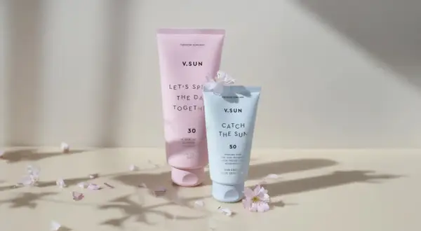 Zwei V.SUN Sonnencreme-Tuben, eine mit LSF 30 für den Körper, die andere mit LSF 50 für das Gesicht, umgeben von zarten Blütenblättern.