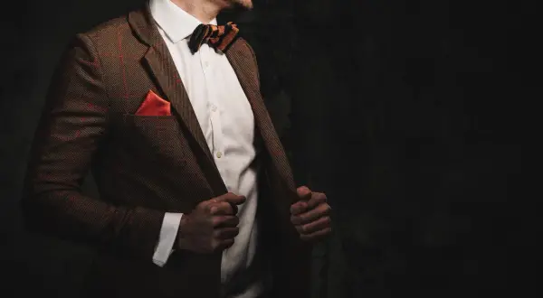 Elegancki mężczyzna w brązowej, tweedowej marynarce w kratkę, białej koszuli i muszce. Stylizacje z tweedową marynarką to klasyka.