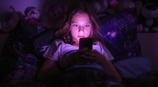 Dziewczynka leżąca w łóżku, patrząca w ekran telefonu w ciemności.