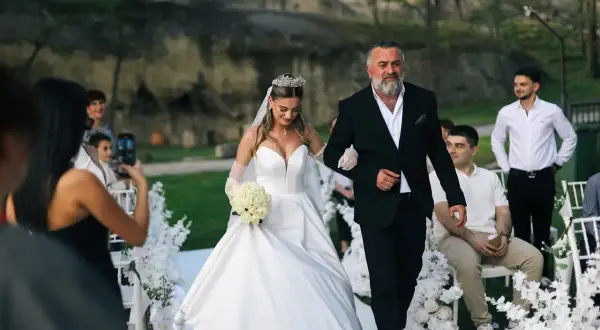 Ojciec prowadzi córkę do ołtarza, wypełniając zasady tradycji. Panna młoda w białej sukni i welonie, z bukietem róż, idzie u jego boku.