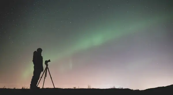 Sylwetka fotografa z aparatem na statywie pod rozgwieżdżonym niebem i zorzą polarną. Efekt bokeh gwiazd dodaje magii.