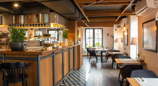Wnętrze restauracji Magiel: drewniany bar, czarne krzesła, stoliki z białymi obrusami i nowoczesne oświetlenie tworzą przytulną atmosferę.