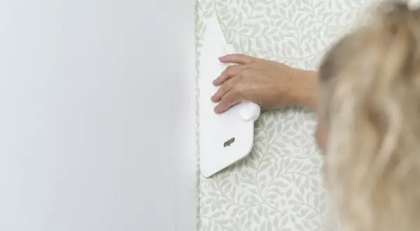 En hand använder en vit spackel för att släta ut en tapetsera non woven med ett ljust bladmotiv.