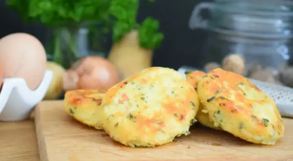 Goldbraune Macaire Kartoffeln, frisch zubereitet, liegen auf einem Holzbrett. Zutaten wie Eier und Petersilie sind im Hintergrund zu sehen.