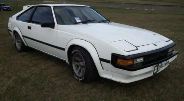 Biała Toyota Supra MK2 z lat 80. stoi na trawie, z widokiem na tor wyścigowy w tle.