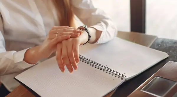 Kobieta sprawdza zegarek, zastanawiając się, ile płatne są nadgodziny. Na stole notatnik i laptop.
