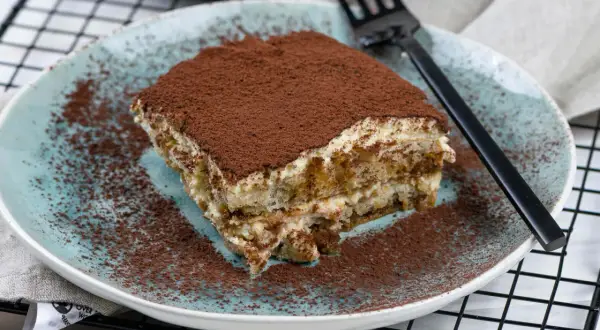 Ein Stück cremiges Tiramisu ohne Alkohol, bestäubt mit Kakaopulver, auf einem blauen Teller.