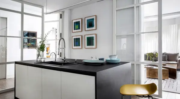 Isla de cocina moderna con encimera oscura, separada del salón por una pared de cristal.
